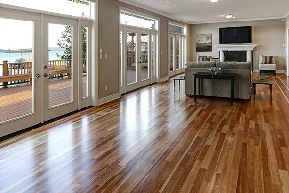 Modern, spacious living room with hardwood flooring, large glass windows, and a kitchen area in the background.
